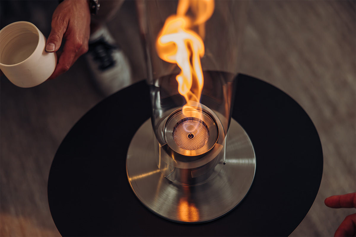 SPIN 900 Bio-Burner mit Eco-Ring und Löschdeckel Eine Person hält den 00696 Spin 1200 Bio-Brenner und platziert ihn in der SPIN Tischfeuer, was der Szene eine funktionale und elegante Note verleiht.