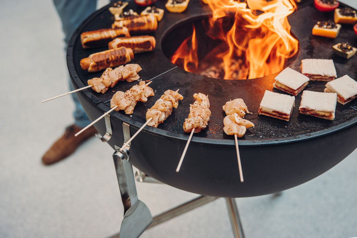 BOWL 70 Fire Bowl with tripod The 00258 Bowl 70 with Tripod integrates seamlessly into the setting, where a person is cooking food on a grill.