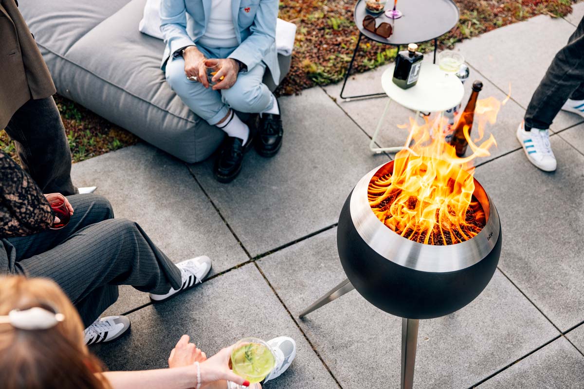 MOON 45 Brasero avec pied haut Un groupe de personnes assis autour d'un feu de camp, avec le 00717 MOON 45 avec un support haut ajoutant une touche fonctionnelle et élégante à la scène.