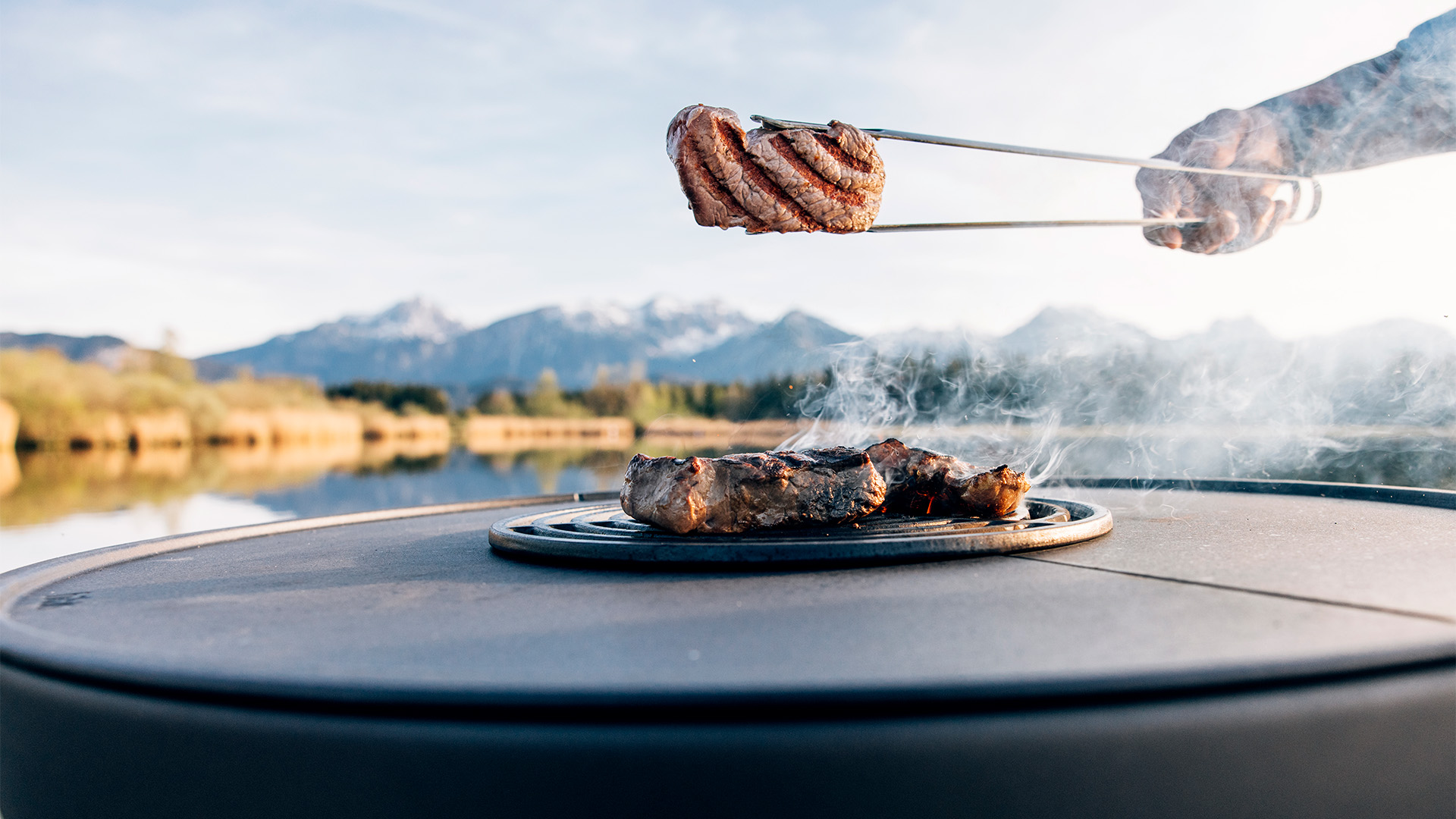 La photo présente une personne en train de Griller de la viande sur un barbecue, avec la Grille de saisie BOWL 70.
