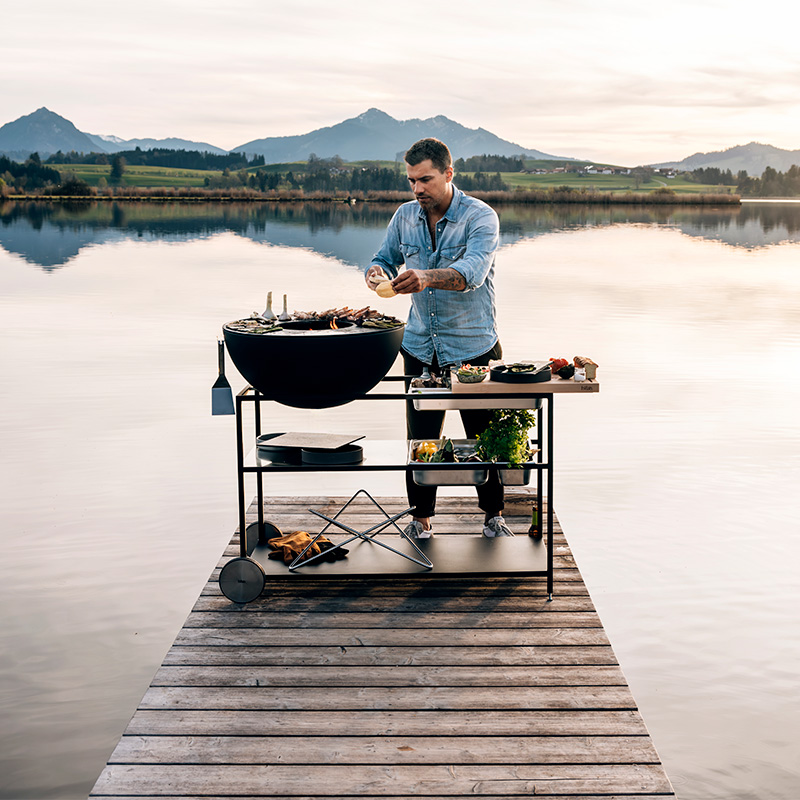 A view of a man sitting on a dock with a grill on it, with the FIRE KITCHEN.
