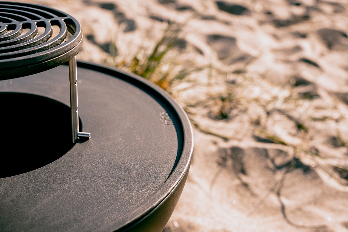 BOWL 57 Sear Grate Abgebildet ist ein schwarzer Feuerkorb auf einem sandigen Strand, mit dem BOWL 57 Wärmekranz.