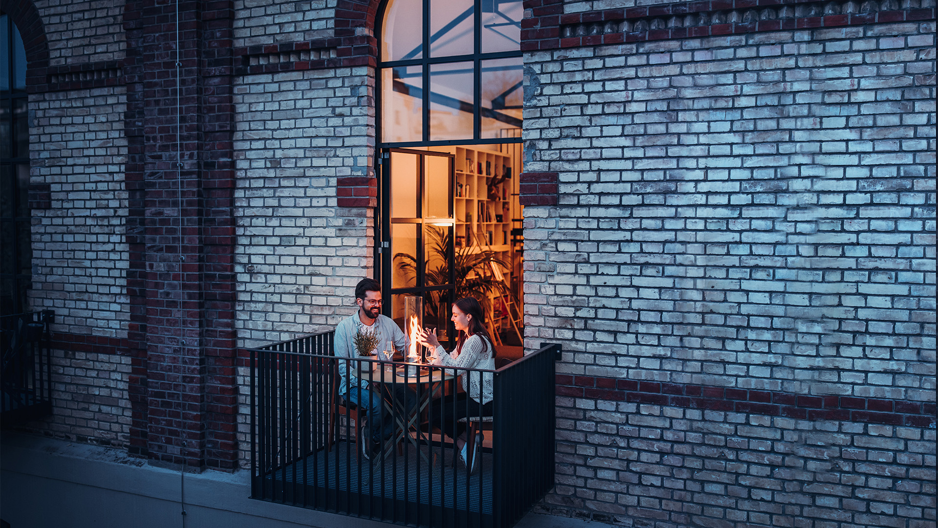 Dargestellt ist ein Mann und eine Frau, die auf einem Balkon sitzen und aus einem Fenster schauen, mit dem SPIN 1200 Tischkamin in Silber.