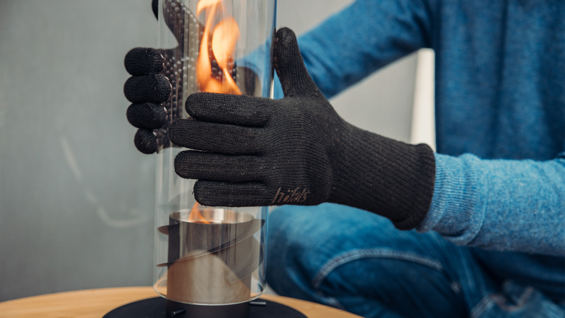 Dieses Bild zeigt eine Person in einem blauen Pullover und schwarzen Handschuhen, die einen Tischkamin mit Feuer in den Händen hält, mit den Fire Gloves Aramid.