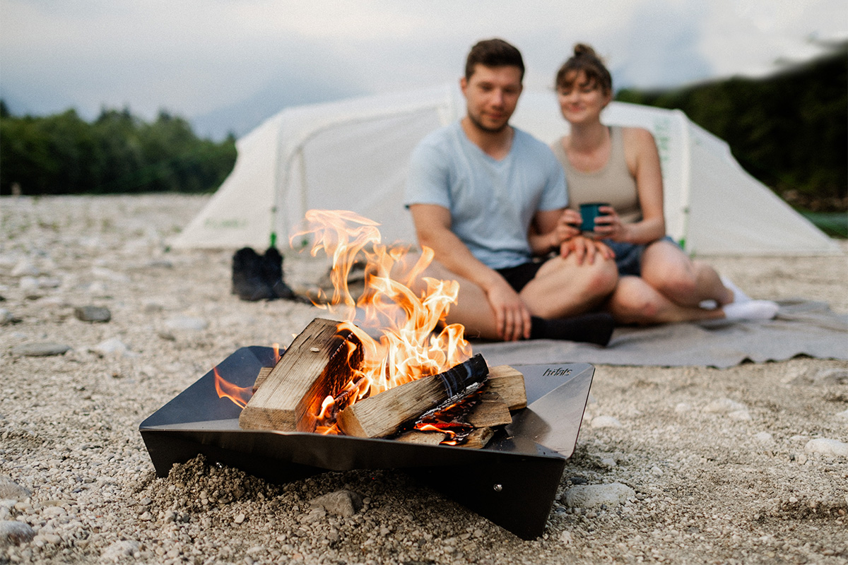TRIPLE 65 Feuerschale Erleben Sie einen Mann und eine Frau, die neben einem Lagerfeuer sitzen, mit dem raffinierten Touch des Triple 65 Kamin.
