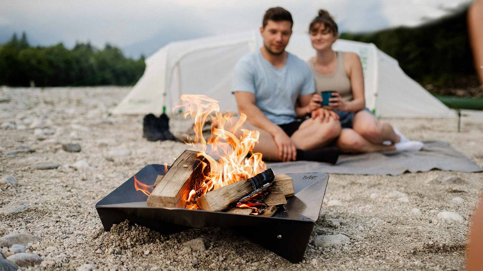 Ein Mann und eine Frau sitzen auf einer Decke am Lagerfeuer, während der Triple 65 Kamin eine funktionale und elegante Note der Szene hinzufügt.