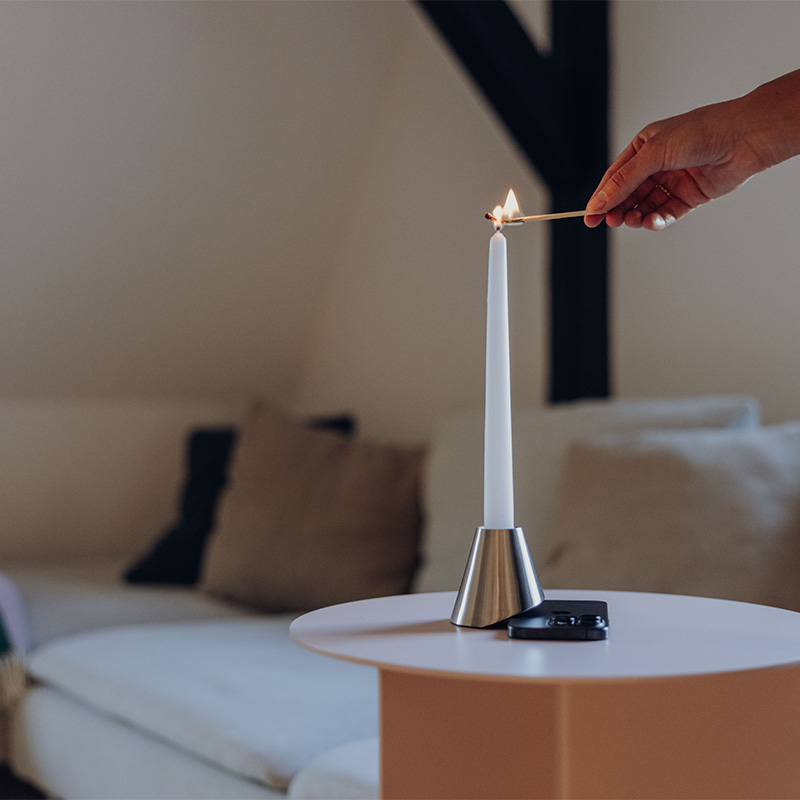 Digi Detox mit Smartphone darunter auf einem kleinen weißen Tisch. Von rechts ist eine Hand sichtbar, die mit einem Streichholz die Kerze anzündet.