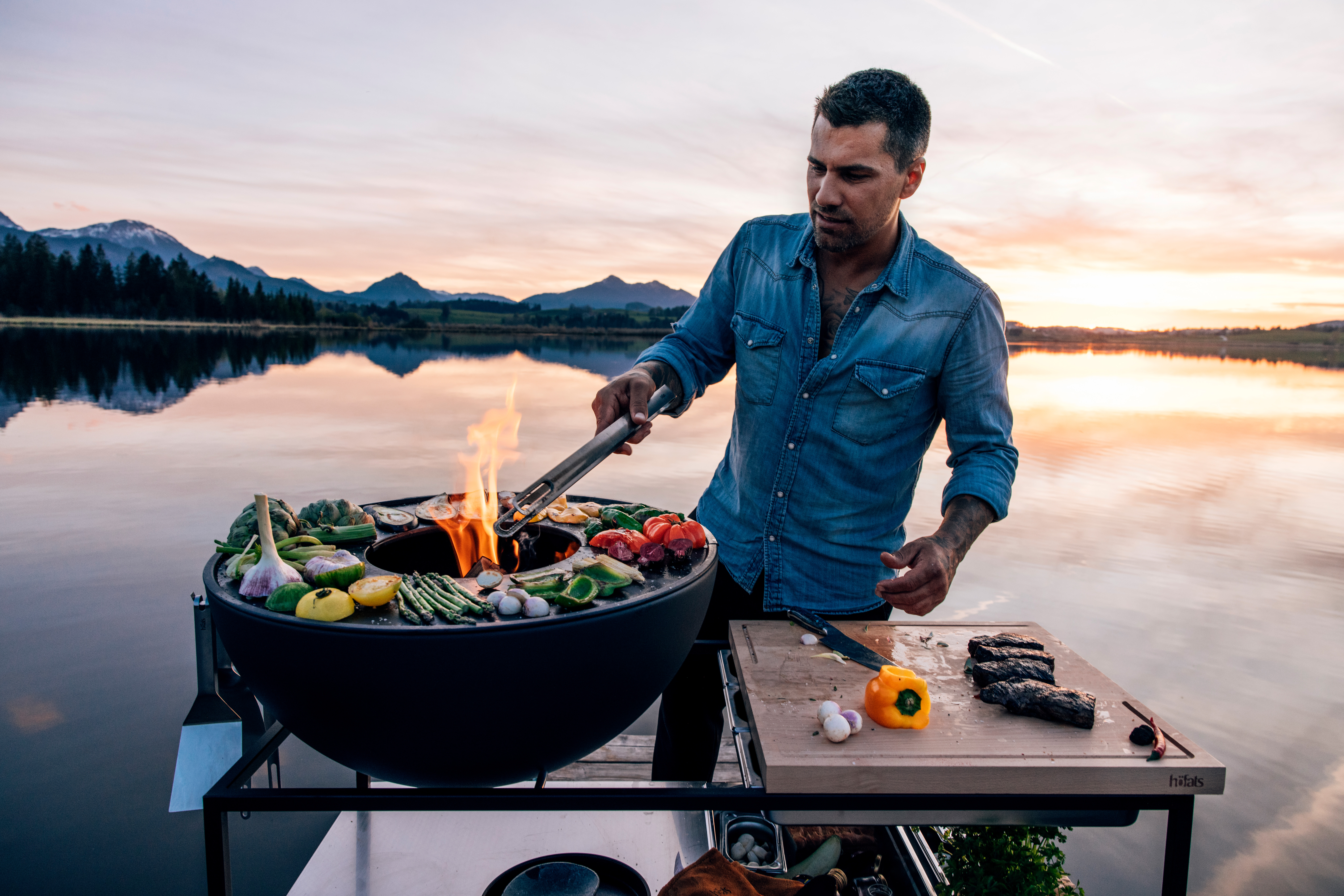 FIRE KITCHEN avec BOWL 70 Set Plancha Grille basse Illustré est un homme qui cuisine sur un gril au bord d'un lac, avec le FIRE KITCHEN avec le BOWL 70 Plancha BBQ Set incl. roues.