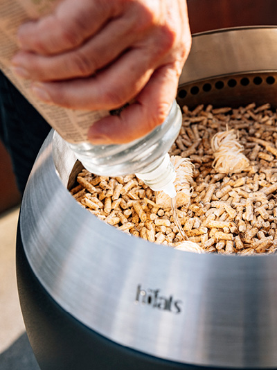 Une personne verse de l'éthanol sur des copeaux de bois dans un Brasero. Montrant comment allumer le MOON 45.