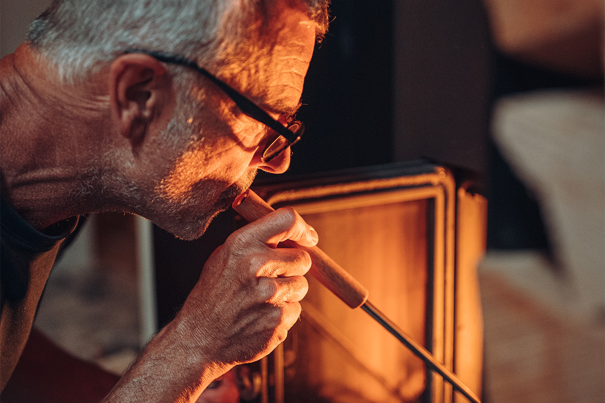Tisonnier avec souffleur de flamme sur pied en inox Le soufflet 00252 avec base en acier inoxydable s'intègre parfaitement dans le décor, où un homme avec des lunettes regarde une cheminée.