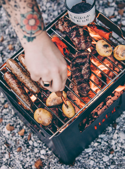 A depiction of a person is cooking meat on a grill, featuring the Beer Box Firebasket as a central and purposeful element.