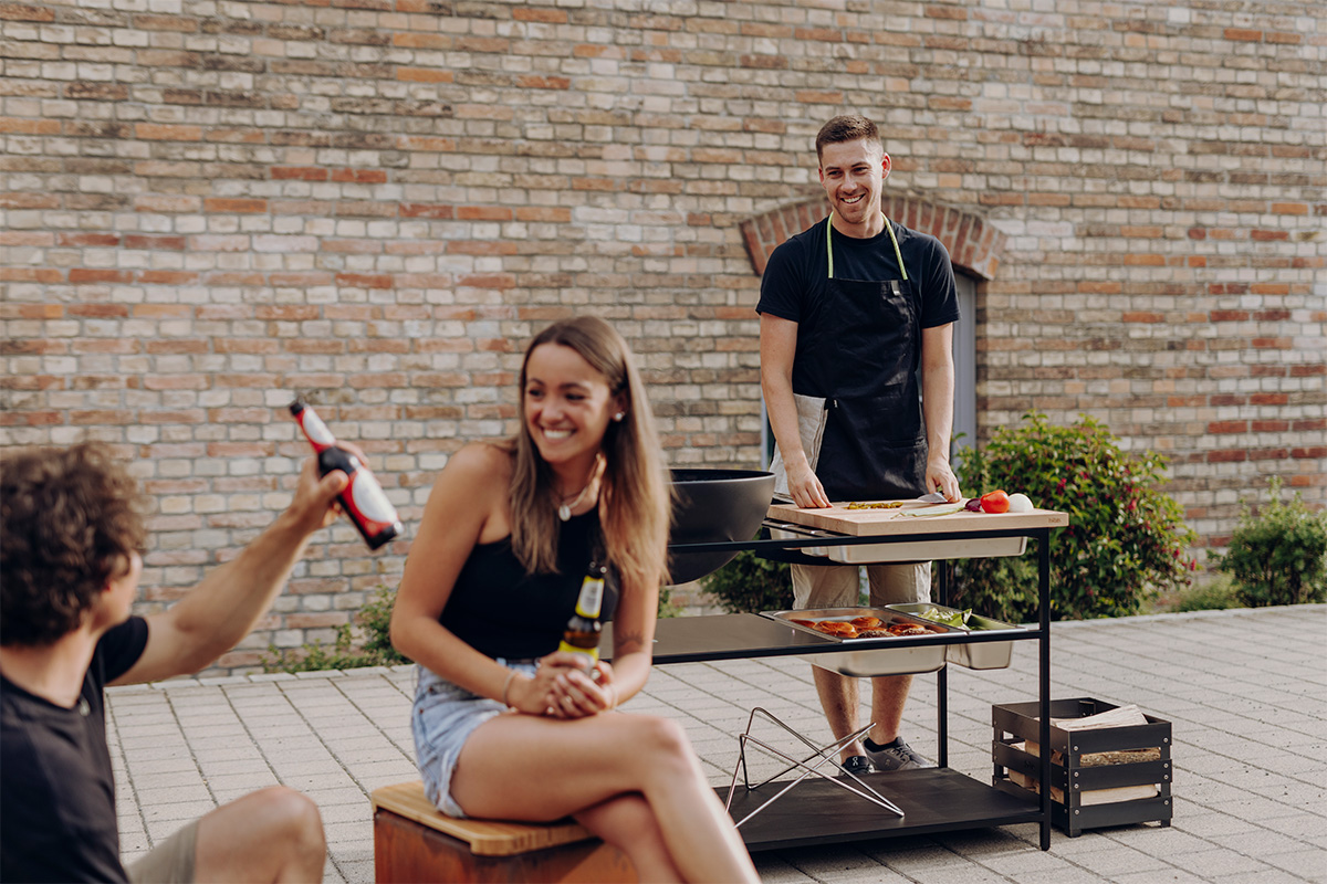FIRE KITCHEN avec BOWL 57 Set Plancha Grille basse La photo présente un homme et une femme assis sur un banc avec un grill, avec le FIRE KITCHEN avec BOWL 57 Plancha BBQ Set incl. roues.