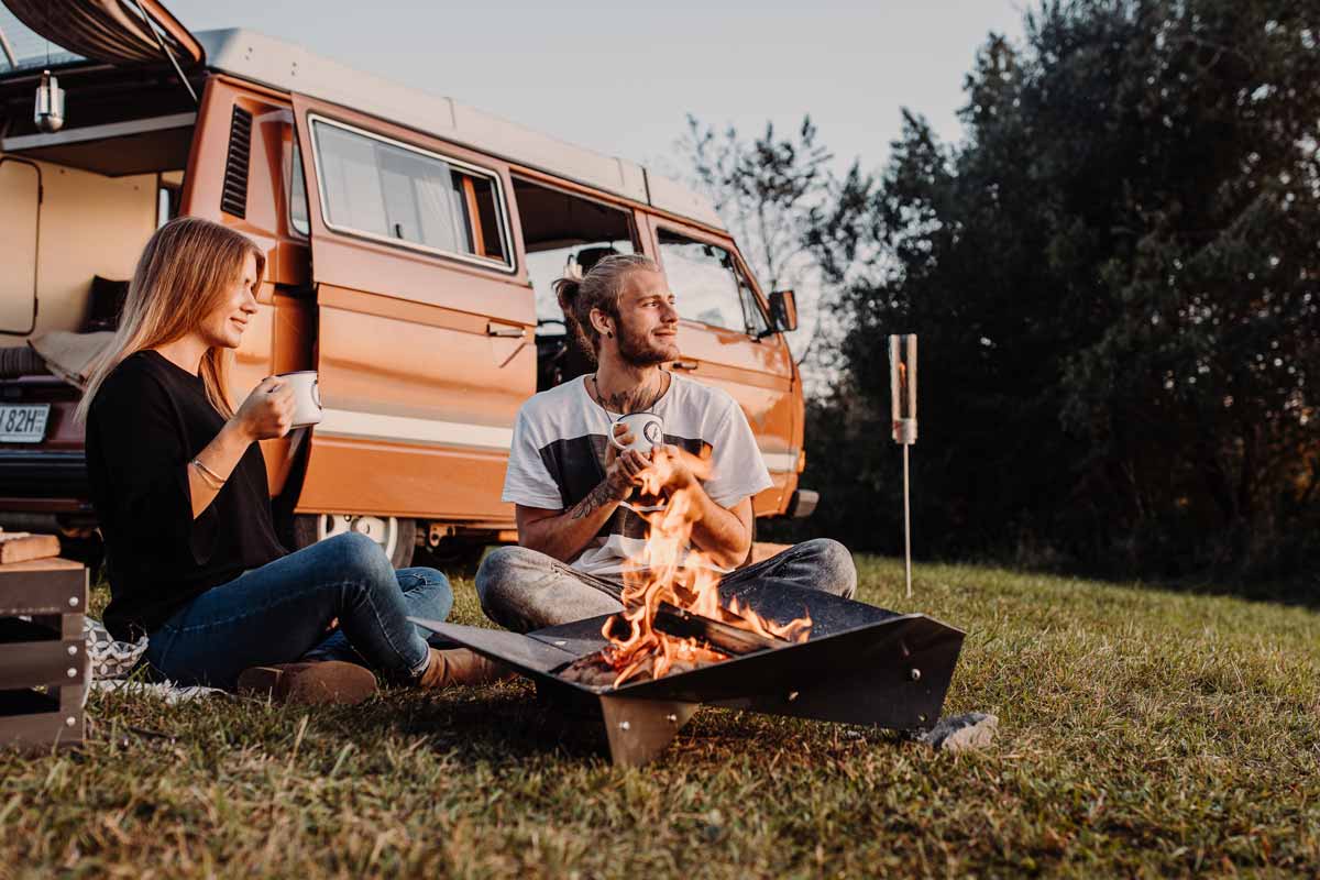 TRIPLE 90 Bol à Feu La photo présente un couple assis sur l'herbe à côté d'un camping-car, avec le TRIPLE 90 Fire BOWL.