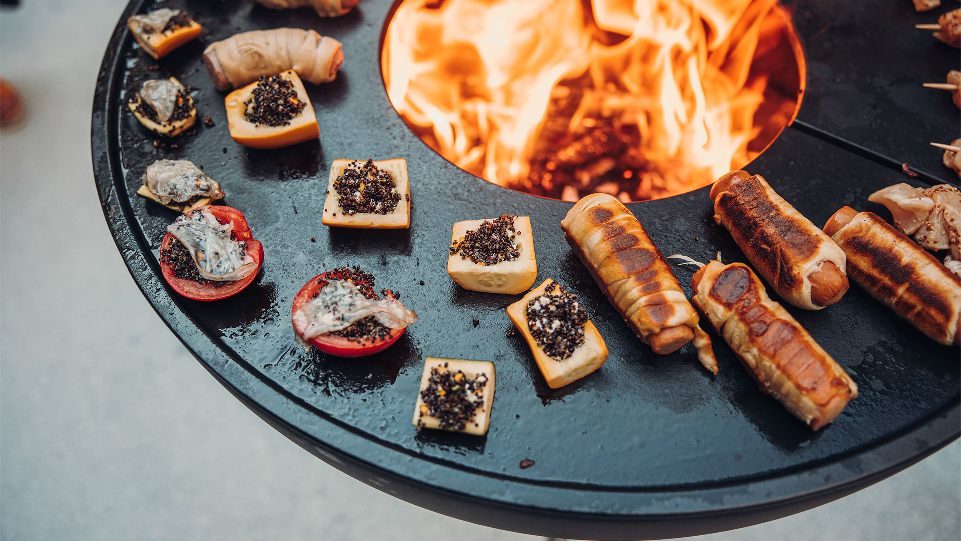 Die 00263 Bowl 70 Plancha fügt sich nahtlos in die Umgebung ein, wo ein Feuerkorb mit einer Menge Fleisch darauf steht.