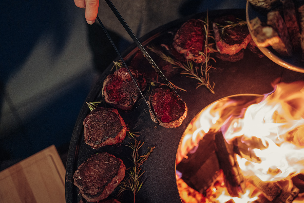 Grillzange schwarz Eine Person grillt Steaks auf einem Grill, bereichert durch die Anwesenheit der schwarzen Zange 00859, die Design und Funktionalität vereint.