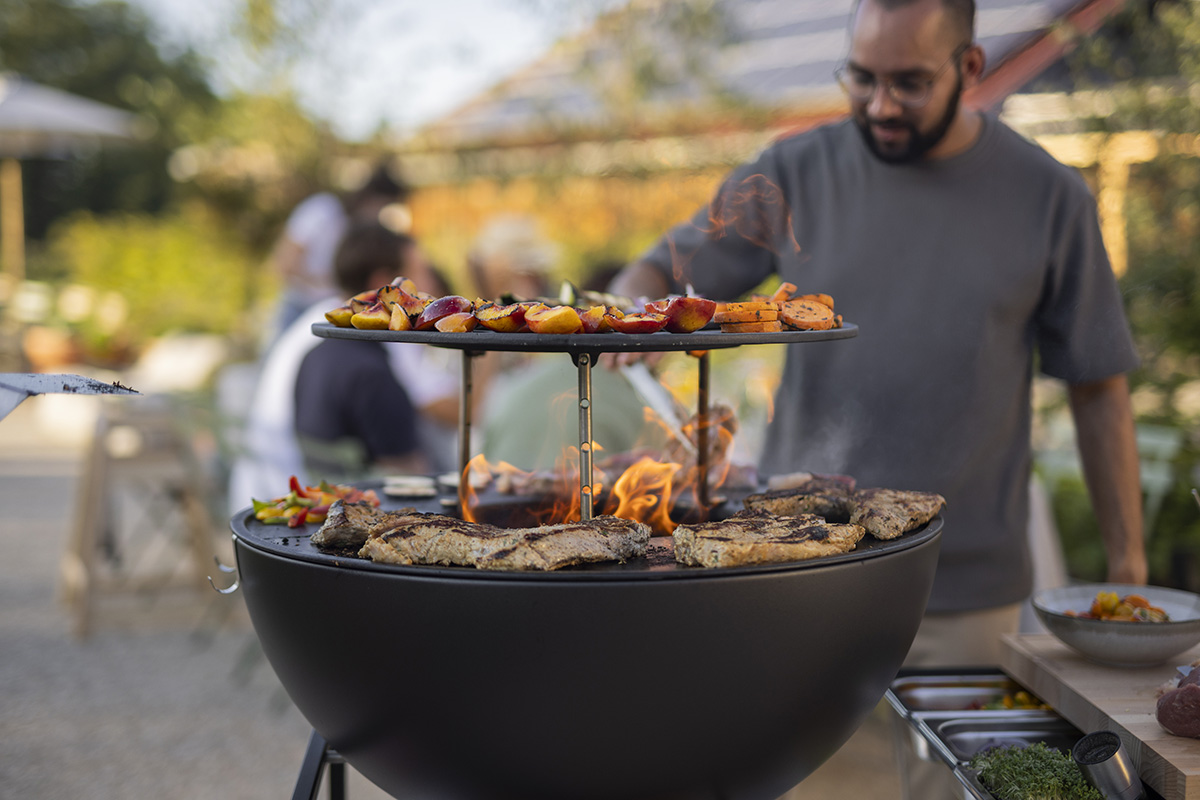 Un homme cuisine de la nourriture sur un gril, avec le BOWL 70 Sear Grate ajoutant une touche fonctionnelle et élégante à la scène.