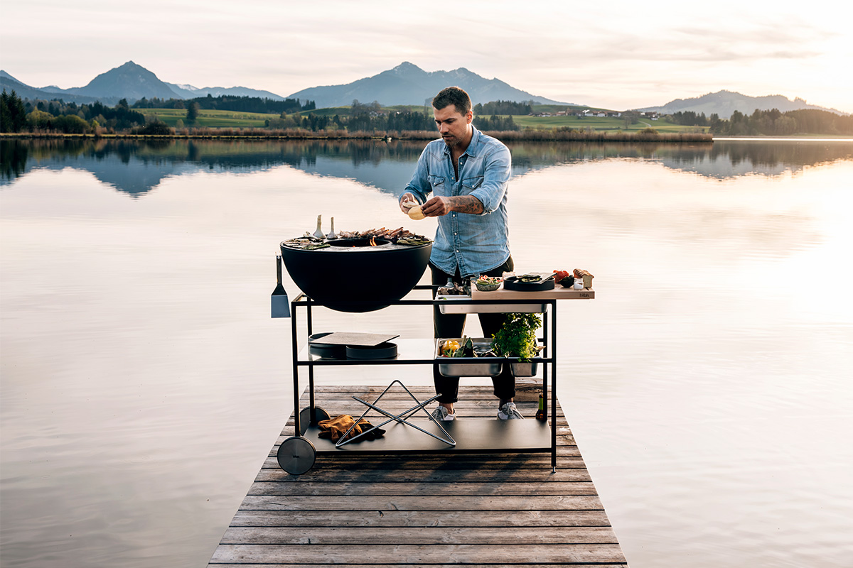 Un homme assis sur un quai avec un grill dessus, enrichi par la présence de l'ensemble de conteneurs en acier inoxydable 00764 FIRE KITCHEN (5 pièces), qui allie design et utilité.