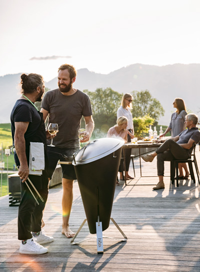 An image showing a group of people sitting at a table on a deck, with the CONE Charcoal Grill.