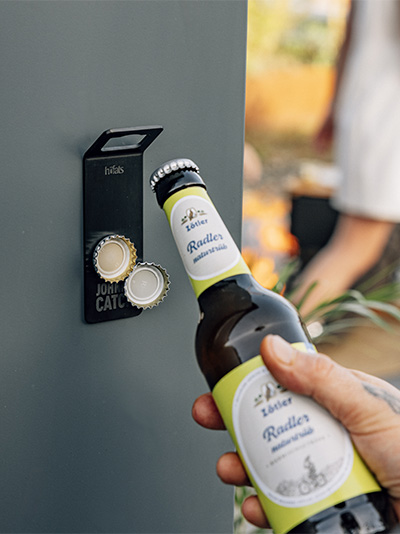 Une personne ouvre une bouteille de bière avec un décapsuleur. L'ouvre-bouteille magnétique Johnny Catch complète le moment avec son design réfléchi.