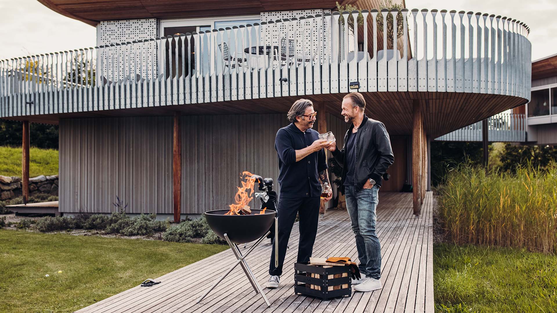 Le BOWL 57 Trépied est présenté avec deux hommes en train de faire un barbecue à l'extérieur d'une maison, créant une image élégante et accessible.