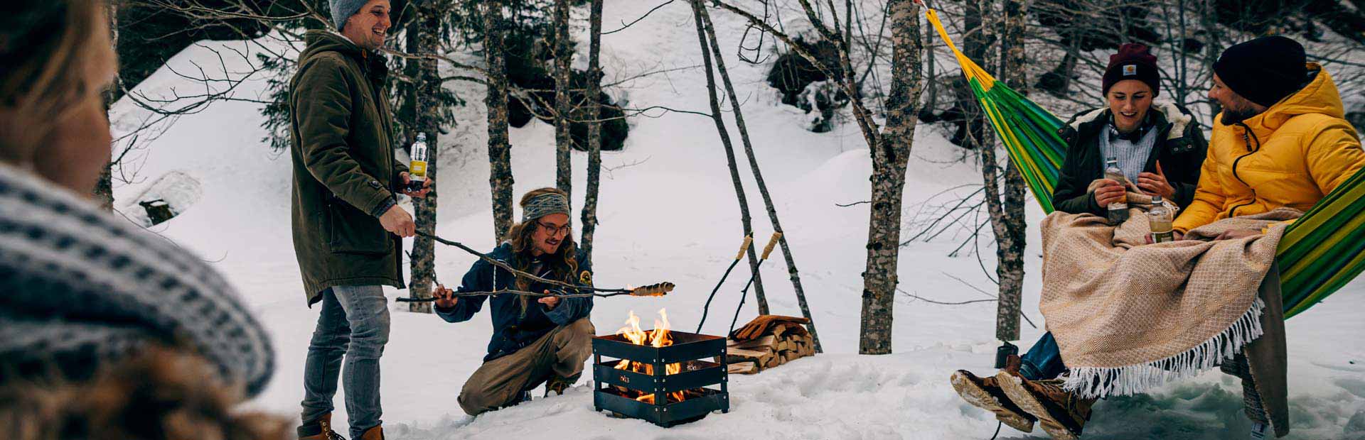 The photo presents people sitting around a campfire in the woods, with the CRATE Fire Basket.