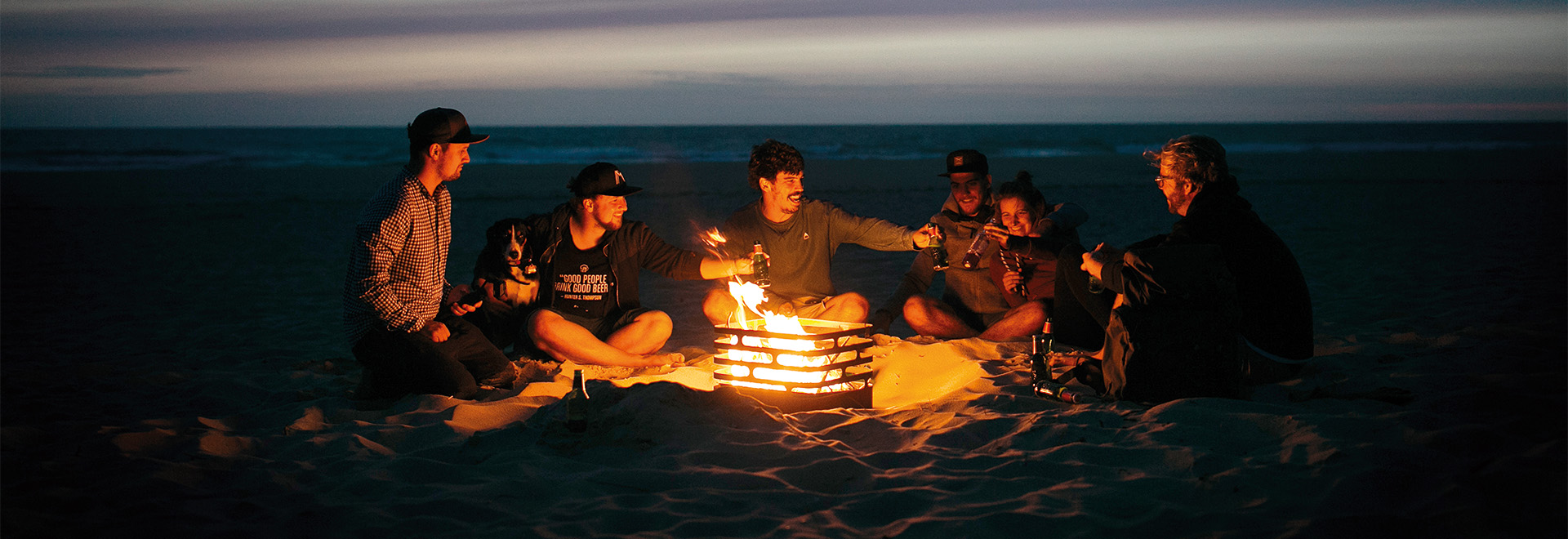 Eine Gruppe von Menschen sitzt um ein Feuer am Strand. Der Cube Feuerkorb ist das zentrale Element der Szene.