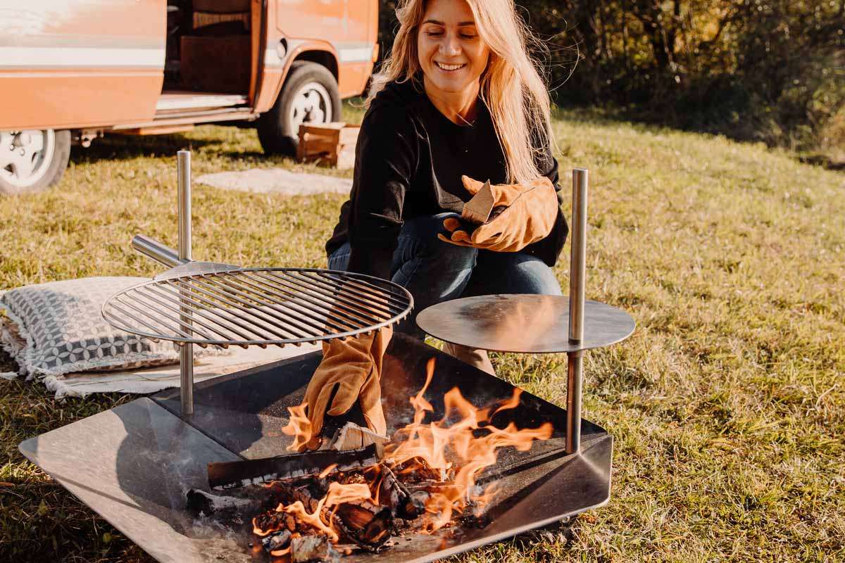 TRIPLE 90 Multi Set The Triple 90 Fireplace Multiset integrates seamlessly into the setting, where a woman sitting on the grass next to a campfire.