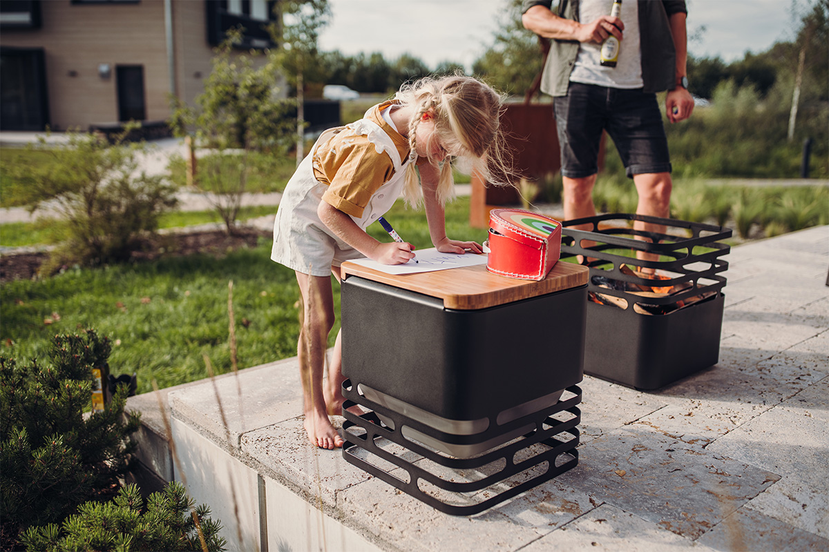Une petite fille est debout sur un bloc de béton, utilisant un CUBE brasero comme support de dessin, enrichi par la présence du CUBE Flip, qui mêle design et utilité.