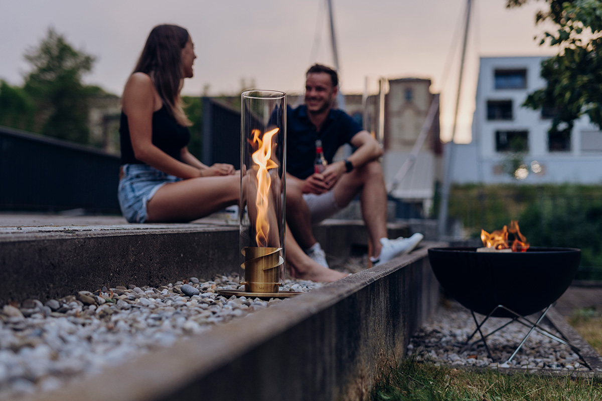 SPIN classic 1200 Cheminée de Table or Une scène accueillante avec un homme et une femme assis sur un banc à côté d'un feu, mis en valeur par la présence du SPIN 1200 Feu de Table or.
