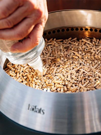 Scene showing a person is pouring liquid ethanol onto wood pellets in a Grill, showing how to light up the MOON 45