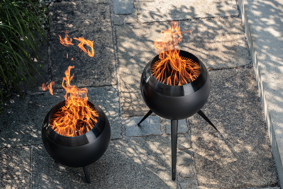 MOON 45 all black Fire Basket with low stand Die MOON mit niedrigem Fuß und MOON mit hohem Fuß sind mit brennender Flamme auf einer Stein Terrasse zu sehen.