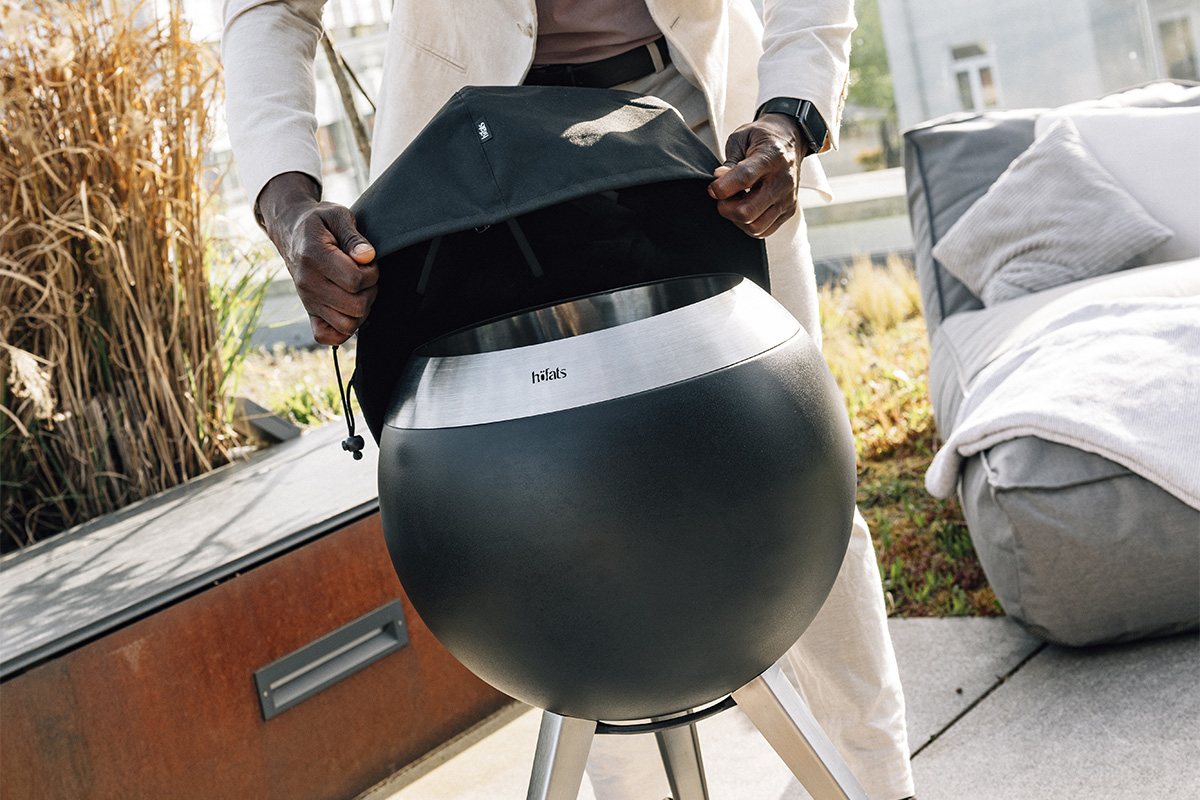 Ein Mann hält eine große schwarze Abdeckung über einem Feuerkorb, wobei die 00815 Moon 45 Abdeckung eine funktionale und elegante Note zur Szene hinzufügt.