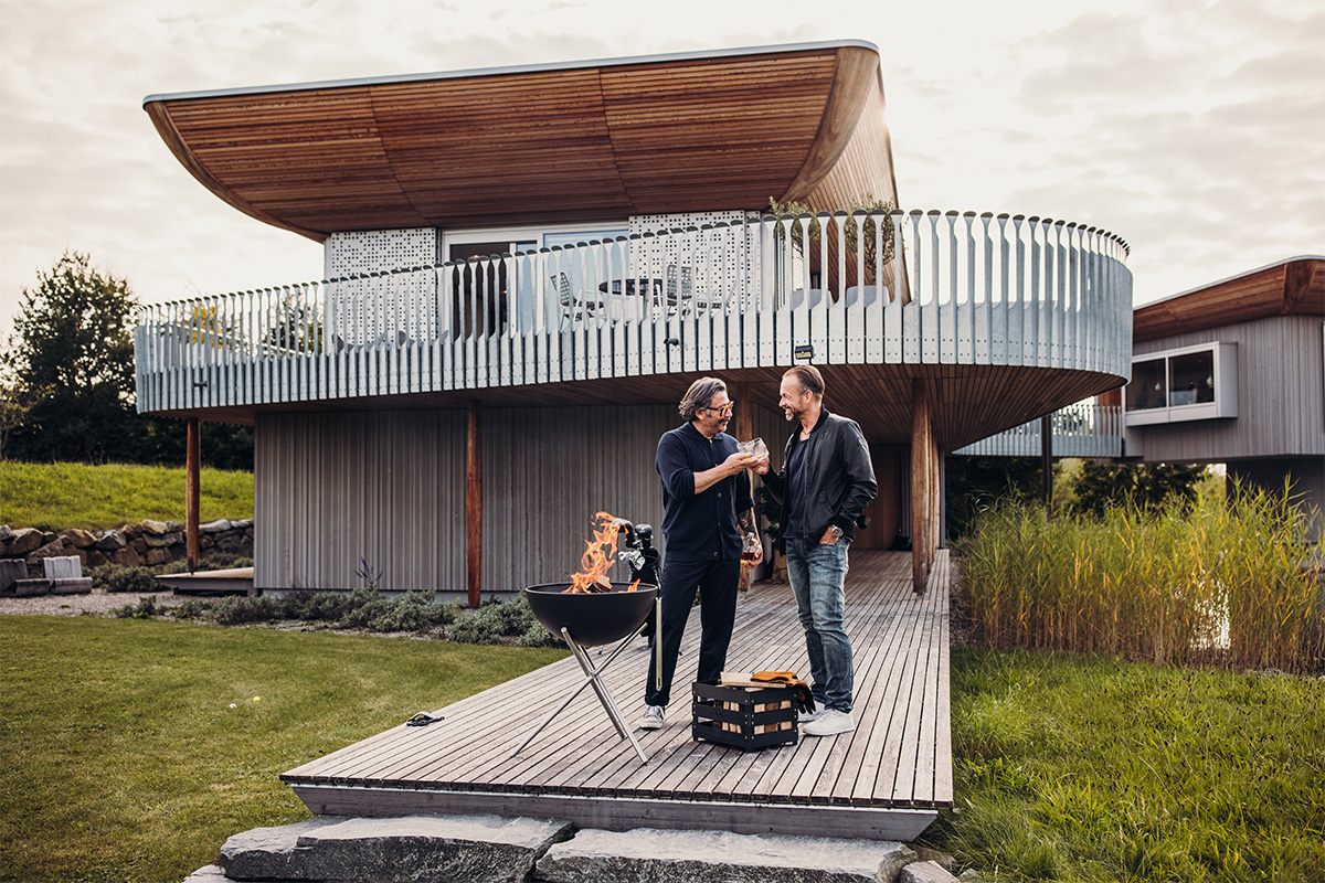 BOWL 57 Feuerschale mit Dreibein Ein Mann steht auf einer hölzernen Terrasse, wobei die 00250 Bowl 57 mit Stativ eine funktionale und elegante Note zur Szene hinzufügt.