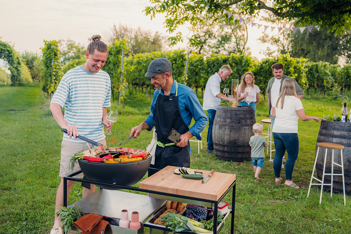 Un groupe de personnes se tenant autour d'un grill, avec le 00739 Fire Kitchen BOWL 57 Plancha Grill Set ajoutant une touche fonctionnelle et élégante à la scène.