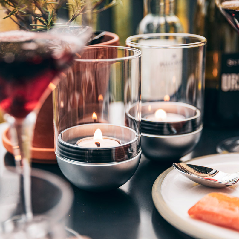 The photo presents a table with a plate of food and a glass of wine, with the LOGO GRAVITY CANDLE M90 Lantern.