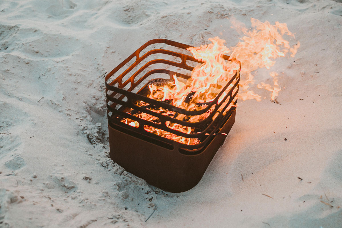 CUBE Brasero acier corten Un brasero est sur la plage avec du sable, avec le CUBE brasero rouillé ajoutant une touche fonctionnelle et élégante à la scène.