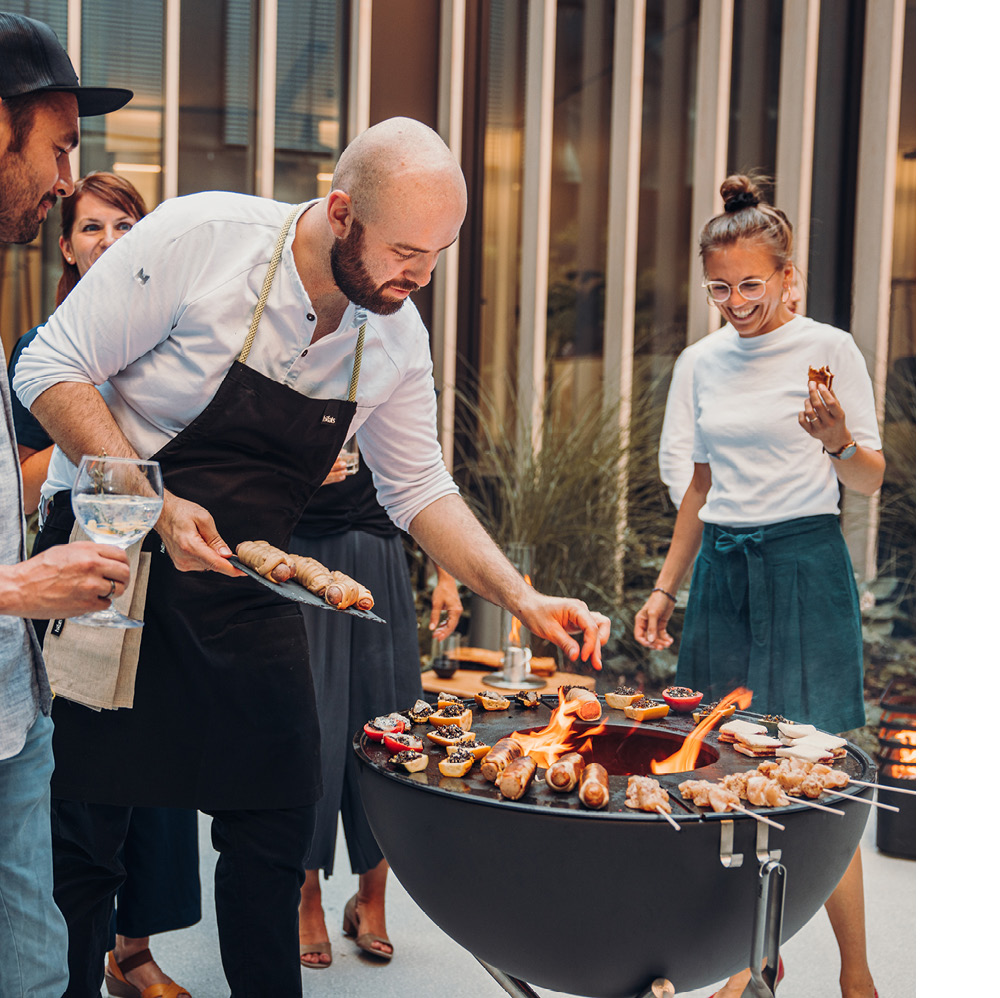 La photo présente un homme en train de cuisiner de la nourriture sur un grill pendant que deux femmes regardent, avec la CUBE Plancha.