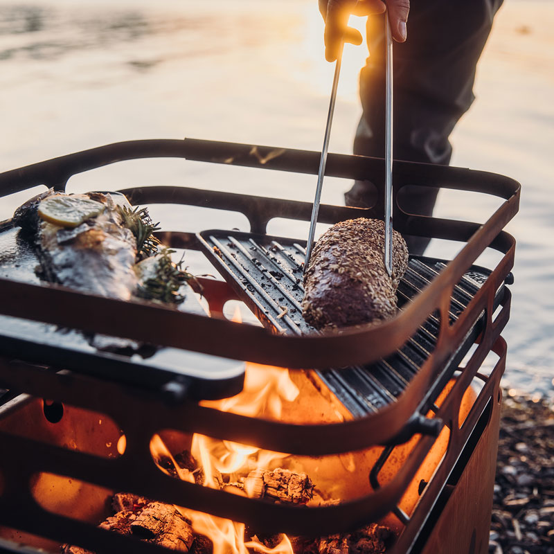 A person is cooking food on a grill the Cube Firebasket complements the moment with its thoughtful design.