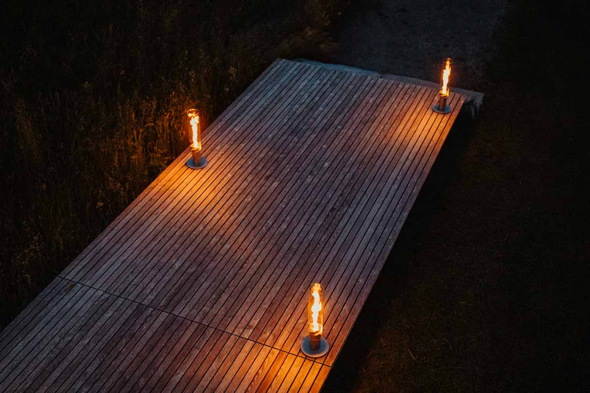 SPIN 900 Stand Elevation silver A view of a wooden deck with two lit candles on it, with the SPIN 90 Table Fire silver.