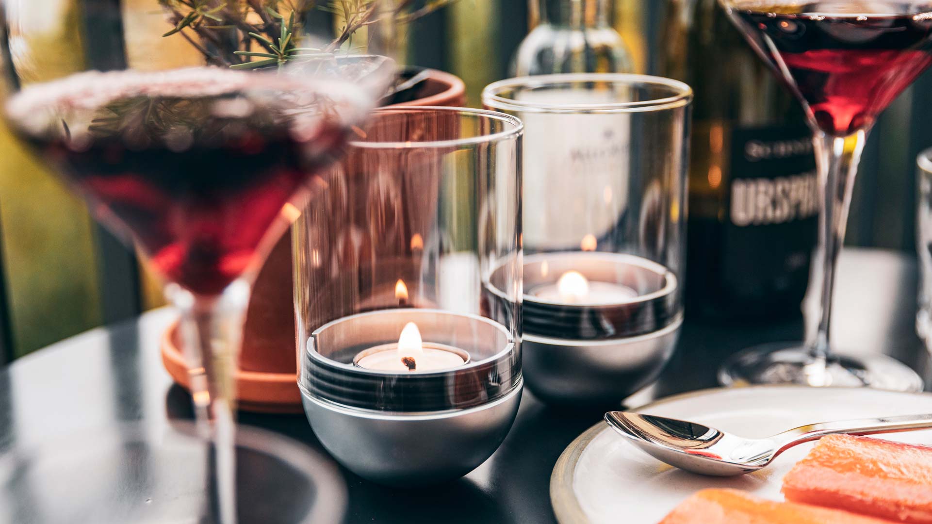 The photo presents a table with a candle, wine glasses and a plate of food, with the LOGO GRAVITY CANDLE M90 Lantern.