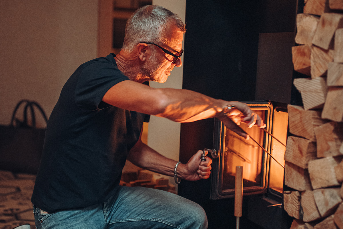 Sur l'image, un homme met le feu dans un poêle à bois, avec des pinces à feu.