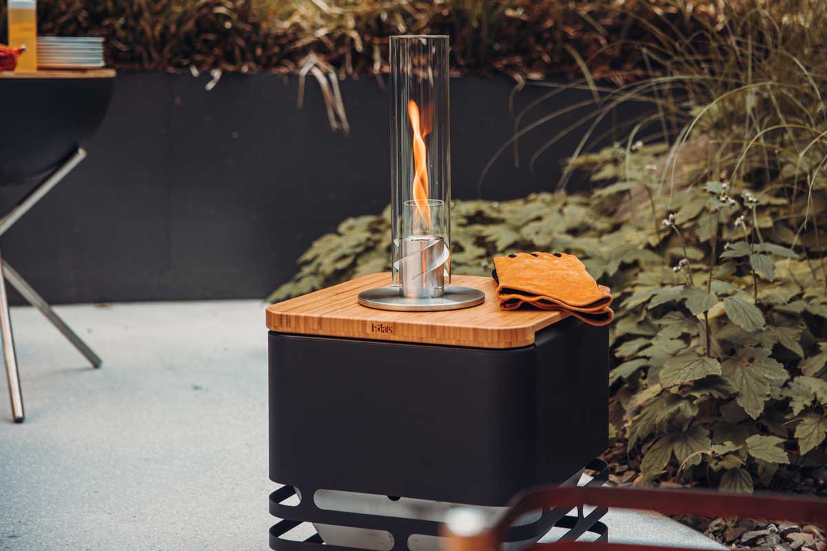 Un gril en métal noir avec une Plancha en bois dessus, tenant le SPIN Feu de Table et des gants en cuir. Montrant l'utilisation de l'ensemble CUBE Flip.