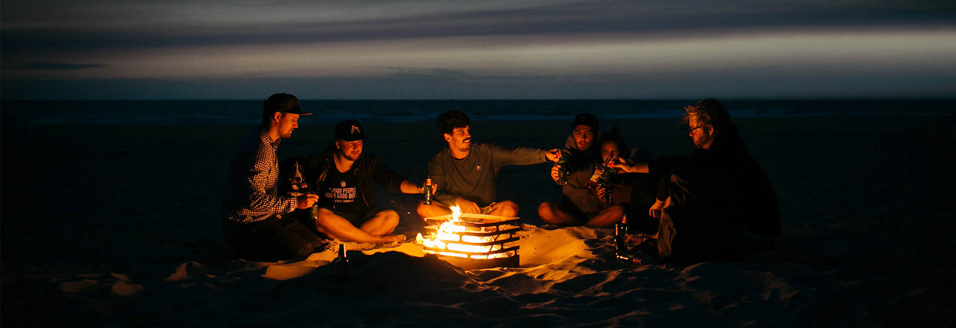 La photo présente un groupe de personnes assises autour d'un feu, avec la cheminée de table logo SPIN.