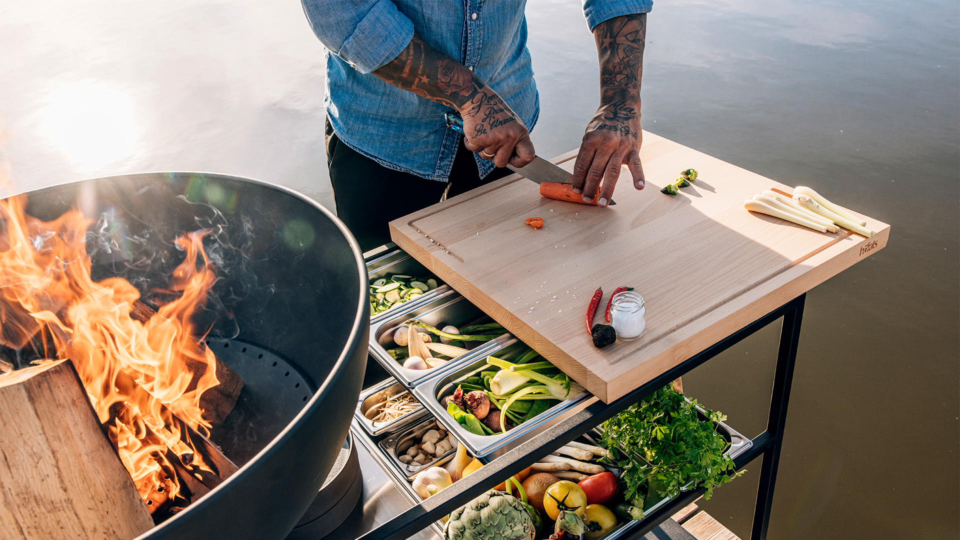 Le plan de travail de cuisine 00614 Fire s'intègre parfaitement dans le décor, où un homme cuisine sur un grill au bord d'un lac.