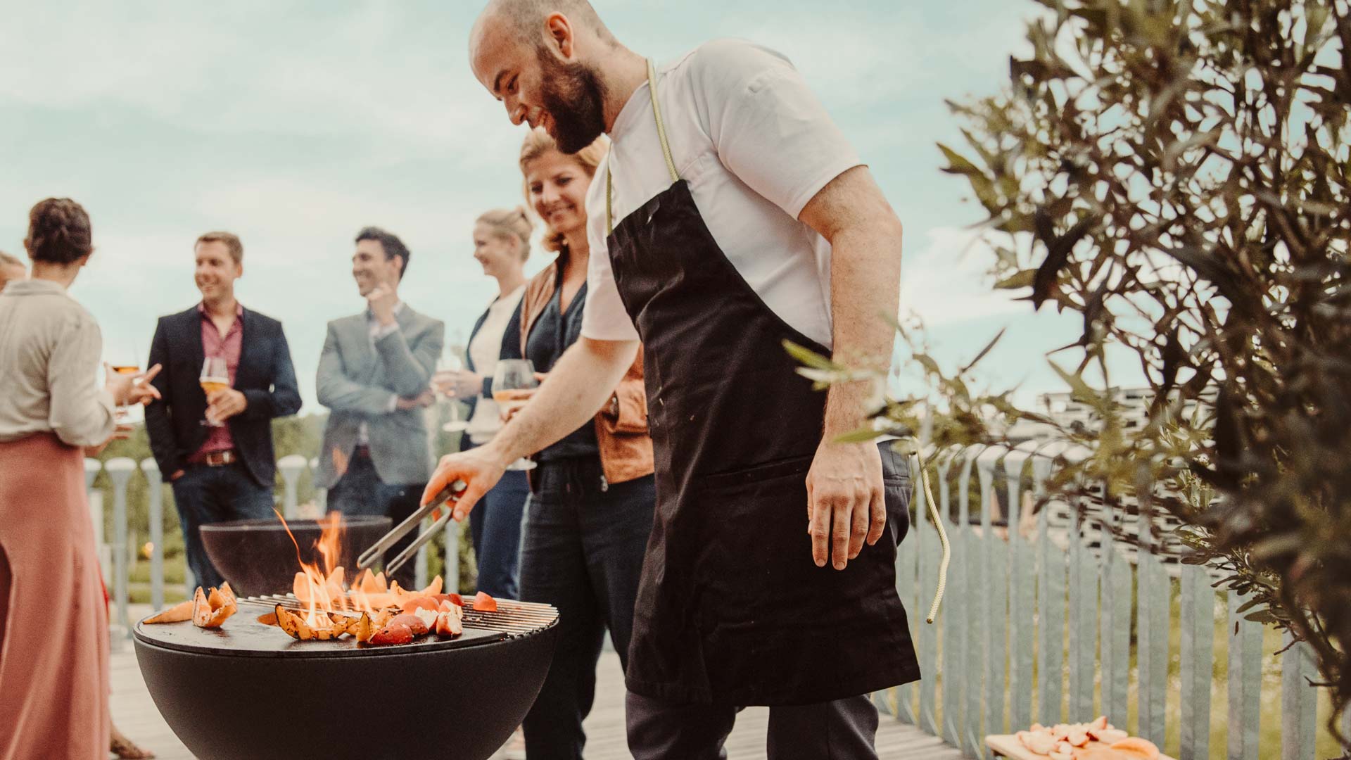 Ein Mann kocht Essen auf einem Grill mit Menschen im Hintergrund.