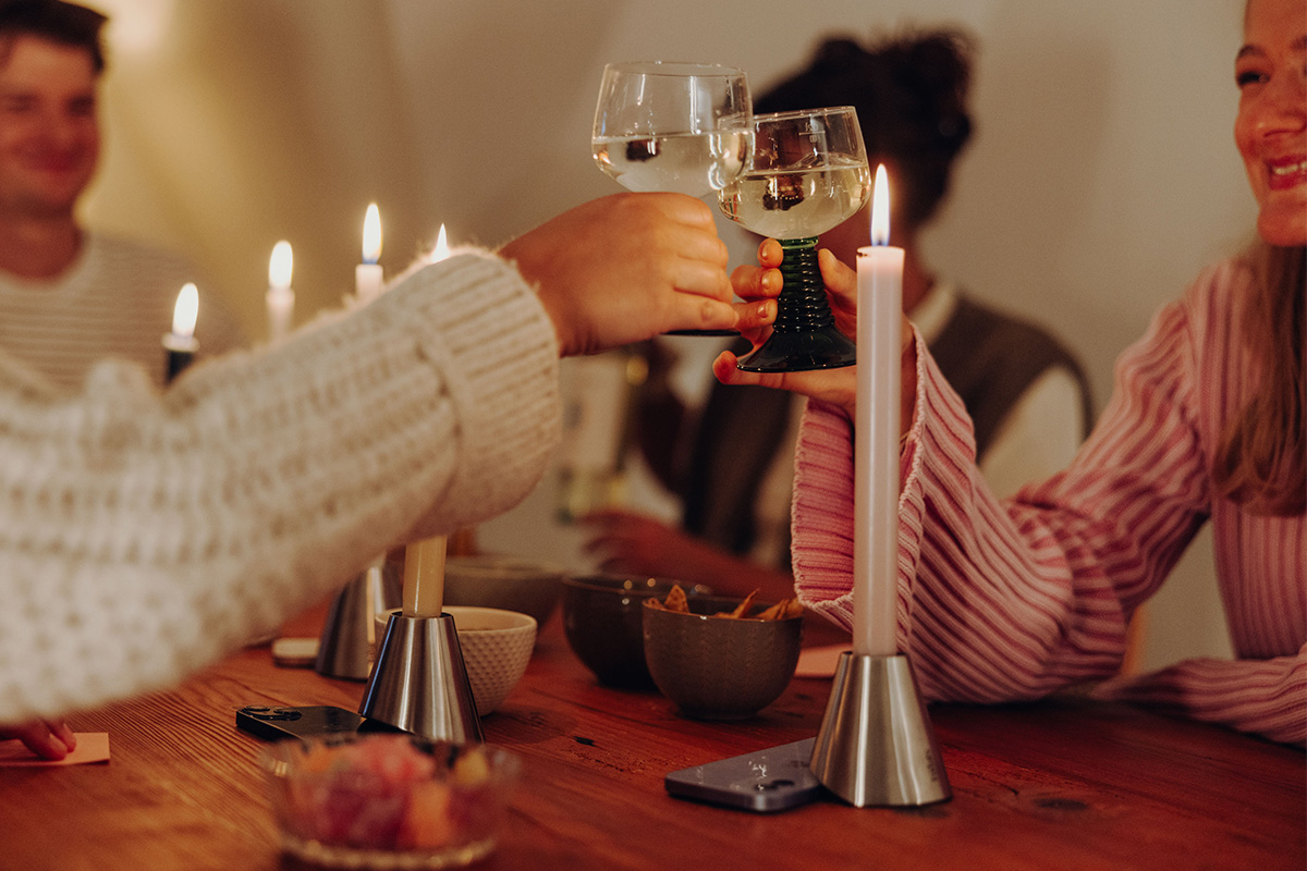 DIGI DETOX Candleholder (Set of 2) Drei Digi Detox mit Smartphone darunter auf Küchentisch. Daneben zwei Hände die mit einem Weinglas anstoßen.