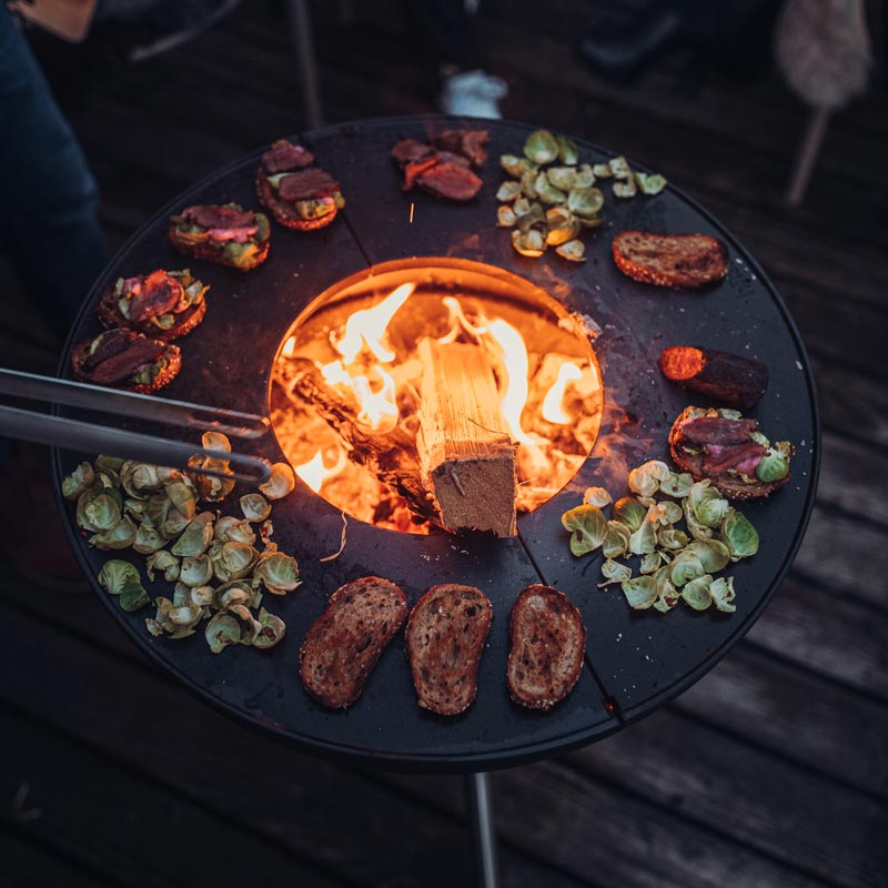 Depicted here is a person roasting sausages over a fire, with the BOWL Fire Bowl.
