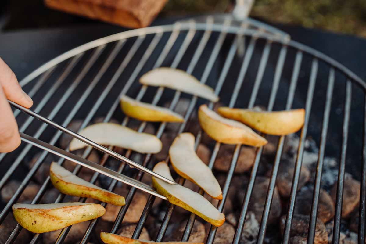 Jemand benutzt eine Zange, um die Kartoffeln vom Grill zu nehmen, während das Triple 65 Kamin BBQ Set eine funktionale und elegante Note zur Szene hinzufügt.