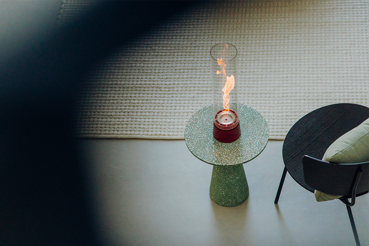 A depiction of a chair and a table fire in a room, featuring the 00814 Spin Air 1200 Tablefire Emaille red as a central and purposeful element.