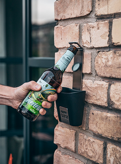 Cette image montre un homme ouvrant une bouteille de bière accrochée au mur avec la tasse JOHNNY CATCH.